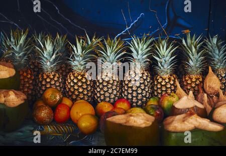 Primo piano di ananas, mele, arance e noci di cocco allineati di fronte a una parete blu Foto Stock