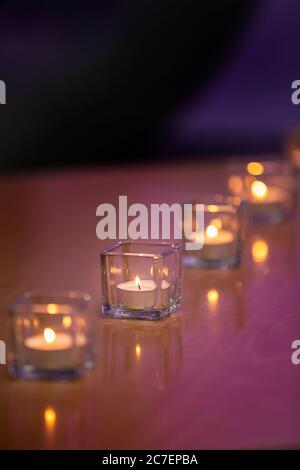 Candele illuminate in contenitori di vetro su un piano da tavolo lucido con sfondo viola Foto Stock