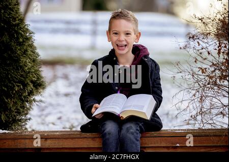 Sole che sorge su un simpatico ragazzino che legge la Bibbia nel mezzo di un parco invernale Foto Stock