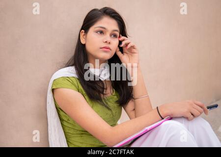 Studente indiano di ragazza teen in ritardo seduto vicino a parete, facendo studio e pensando mentre leggendo sul libro in esterno all'ora di giorno. Foto Stock