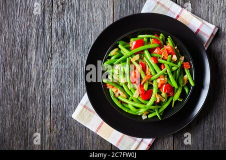 Frittura di fagioli verdi con cipolle e pomodori, Fry bodi in una ciotola nera su un tavolo di legno, vista panoramica dall'alto, piatto, spazio libero Foto Stock