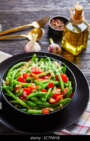 Frittura di fagioli verdi con cipolle e pomodori, Fry bodi in una ciotola nera su un tavolo di legno con posate dorate, vista panoramica dall'alto Foto Stock