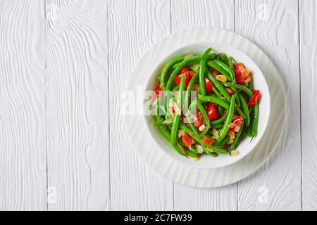 Frittura di fagioli verdi con cipolle e pomodori, frittura bodi in una ciotola bianca su un tavolo di legno, vista panoramica dall'alto, piatto, spazio libero Foto Stock