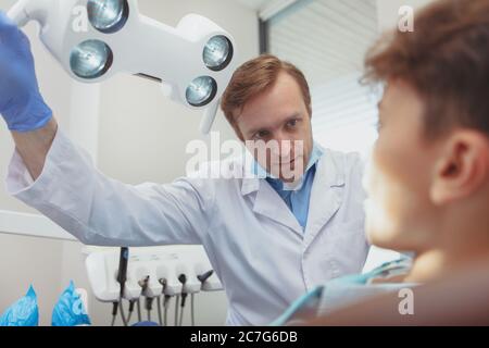 Cure mediche, concetto di odontoiatria. Dentista professionista maturo che regola la lampada dentale prima di esaminare il suo piccolo paziente. Raccordo a T di controllo dentista maschio Foto Stock