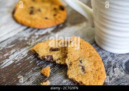 Vista a fuoco superficiale di biscotti al cioccolato preparati di fresco, parzialmente mangiati visti su una superficie di cucina in legno insieme ad una tazza di caffè. Foto Stock