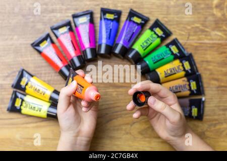 ZAGABRIA, CROAZIA - 17 dicembre 2019: Mani del bambino che tengono la vernice acrilica arancione sopra il vecchio tavolo di legno marrone invecchiato e vernice acrilica colorata. Foto Stock