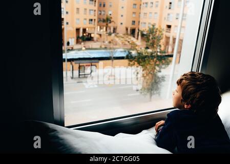 Triste e malinconico ragazzo guarda attraverso una finestra nel suo appartamento durante il suo confinamento Foto Stock