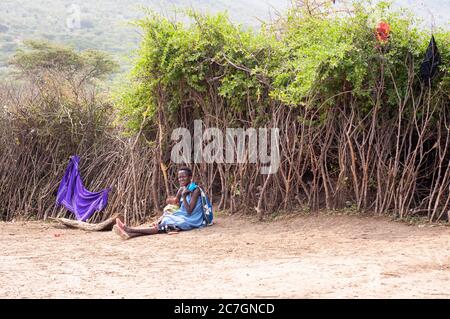 Madre e bambino Maasai, con abbigliamento tradizionale seduto sul pavimento, in un villaggio maasai, nella Riserva Nazionale di Maasai Mara. Kenya. Africa. Foto Stock