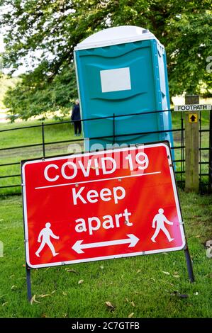Un cartello di avvertimento sociale Covid-19 in occasione di un evento all'aperto con un wc portatile in plastica sullo sfondo. Foto Stock