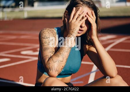 Attraente atleta bagnata in abbigliamento sportivo che copre il viso con le mani mentre si riposa dopo l'allenamento allo stadio cittadino Foto Stock