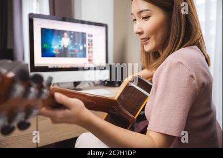 Donna asiatica chitarrista e vlogger che suona la chitarra nel computer anteriore a casa, mentre la distanza sociale di coronavirus focolaio Foto Stock