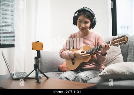 Blogger donna asiatica che registra e suona la chitarra dal vivo sui social media. Concetto di lezione online di chitarrista mentre si è a casa Foto Stock