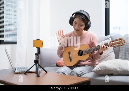 Blogger donna asiatica che registra e suona la chitarra dal vivo sui social media. Concetto di lezione online di chitarrista mentre si è a casa Foto Stock