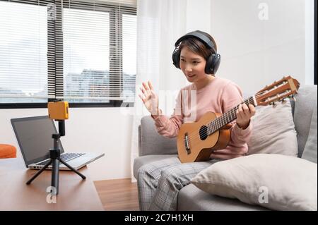 Blogger donna asiatica che registra e suona la chitarra dal vivo sui social media. Concetto di lezione online di chitarrista mentre si è a casa Foto Stock