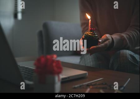 Donna asiatica che festeggia il compleanno di un collega con una videochiamata virtuale a casa mentre è isolata, resta a casa. Torta della tazza con la mano si accende Foto Stock