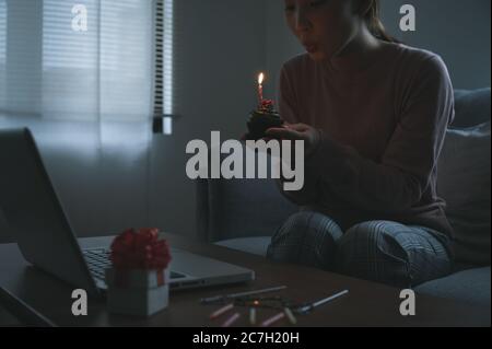 Donna asiatica che festeggia il compleanno di un collega con una videochiamata virtuale a casa mentre è isolata, resta a casa. Torta della tazza con la mano si accende Foto Stock