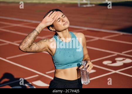 Ragazza atleta sconvolta in top sportivo con una bottiglia d'acqua che ricopre con cura la mano vicino alla testa dopo l'allenamento allo stadio Foto Stock