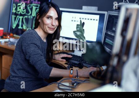 Carino giovane donna Video Editor Freelancer con capelli scuri funziona sul computer mentre si siede al tavolo. L'artista Girl sta elaborando il video Foto Stock