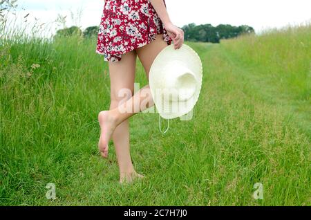 Ragazza a piedi nudi che cammina su erba verde. Donna con gonna colorata, che tiene in mano il cappello estivo. Estate in Polonia, bella campagna in bassa Slesia. Foto Stock