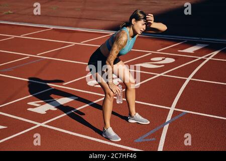 Giovane atleta in abbigliamento sportivo con una bottiglia d'acqua stancata in ginocchio dopo aver corso allo stadio Foto Stock