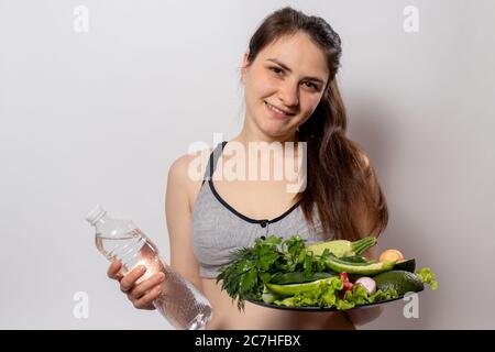 La ragazza tiene una bottiglia d'acqua e un piatto con verdure verdi nelle mani. Nutrizione adeguata, verdure nella dieta, dieta del keto, disintossicazione del corpo Foto Stock