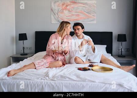 Giovane uomo brunetta in accappatoio bianco e bella donna bionda in rosa velevt accappatoio sognante parlando con tazze di caffè in mani. Bella coppia che ha Foto Stock