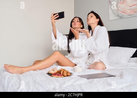 Due belle donne in accappatoi bianchi che inviano felicemente baci aria scattando foto insieme sul cellulare. Le ragazze graziose che hanno la prima colazione a letto con Foto Stock