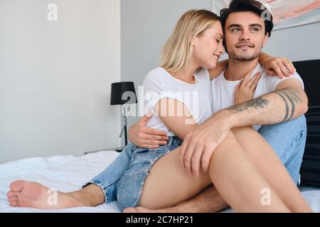 Attraente uomo bruna sorridente che guarda da parte sognante mentre bella donna bionda lo abbracciava felice. Giovane bella coppia in magliette bianche seduta Foto Stock