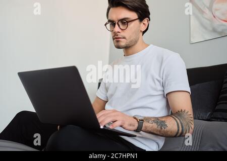 Giovane e attraente uomo di brunetta in T-shirt e occhiali seduta sul letto a casa moderna e con cura di utilizzare sul computer portatile Foto Stock