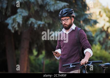 Moto millennial commuter in casco passeggiate con un telefono. Pedalando in città, trasporto verde, uso della bicicletta per girare la città Foto Stock