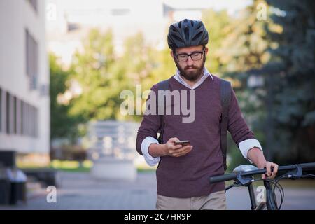 Moto millennial commuter in casco passeggiate con un telefono. Pedalando in città, trasporto verde, uso della bicicletta per girare la città Foto Stock