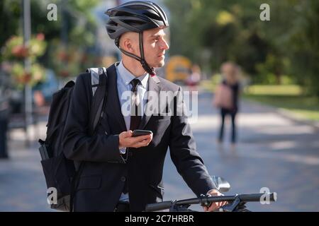 Ritratto di un commuter maschile in un vestito e casco da bicicletta con un telefono. Pedalando per la città, andando a lavorare in ufficio in bicicletta Foto Stock