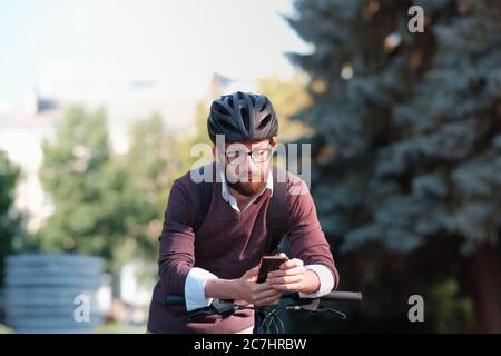 Moto millennial commuter in casco con un telefono. Pedalando in città, trasporto verde, uso della bicicletta per girare la città Foto Stock