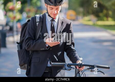 Commuter maschile in bicicletta in un vestito e casco con un telefono. Pedalando per la città, andando a lavorare in ufficio in bicicletta Foto Stock