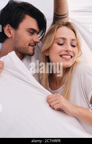 Giovane attraente uomo bruna sognante guardando bella donna bionda sorridente che giace insieme sotto coperta nel letto a casa Foto Stock