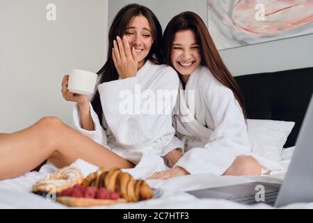 Due belle donne in accappatoi bianchi che guardano i film sul computer portatile insieme a deliziosi pasticcini e caffè. Ragazze sorridenti graziose che hanno Foto Stock