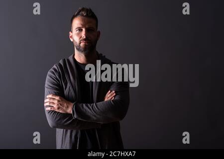 Un grave bel giovane in una felpa con cappuccio grigio in piedi di fronte a uno sfondo grigio scuro in un studio. Foto Stock