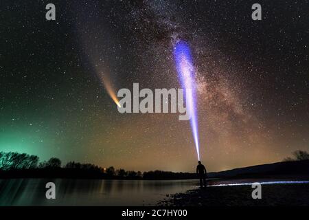 Silhouette di un uomo con torcia da testa in piedi sulla riva del fiume che illumina un fascio di luce sulla galassia di Milkyway e la cometa di Neoswise con coda chiara nel buio Foto Stock