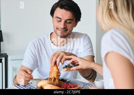 Giovane attraente bruna sorridente uomo in T-shirt bianca seduta al tavolo con la ragazza che ha una deliziosa colazione in casa moderna accogliente Foto Stock