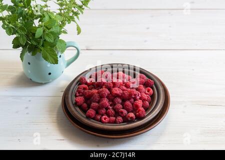 Lamponi freschi maturi rossi in un piatto marrone su un tavolo di legno bianco. Un mazzo di balsamo al limone in una tazza da tè. Delizioso dessert estivo Foto Stock