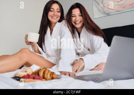 Due belle donne allegre in accappatoi bianchi che guardano con gioia film su computer portatile insieme a deliziosi dolci e caffè. Ragazze graziose che hanno Foto Stock