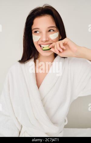 Giovane bella donna sorridente in accappatoio bianco con maschera cosmetica sul viso seduta sul letto mangiare fetta di cetriolo felicemente guardando da parte in moderno Foto Stock