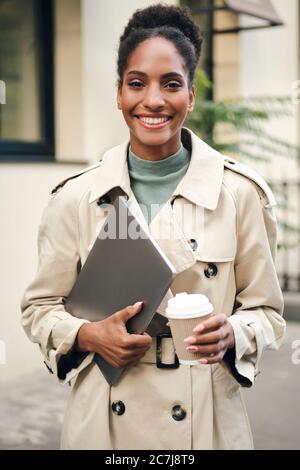 Allegra donna d'affari afroamericana in trench elegante cappotto con computer portatile e caffè per andare gioiosamente guardando in macchina fotografica all'aperto Foto Stock