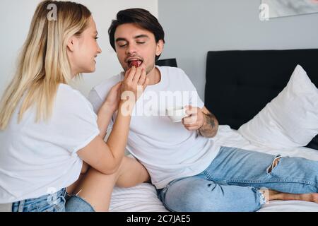 Giovane uomo bruna che tiene in mano una tazza di caffè mangiando in modo sognante la fragola mentre bella donna bionda lo nutrono. Bella coppia in T-shirt bianca Foto Stock