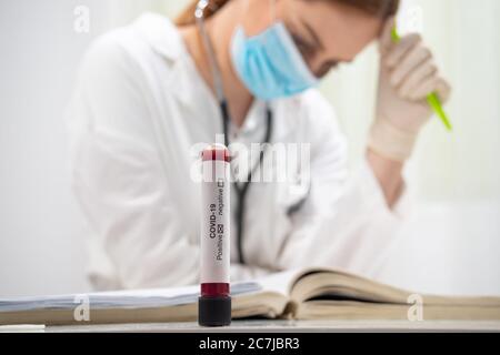 Provetta per analisi del sangue per virus corona positivo, e medico femminile con maschera stanca in background Foto Stock