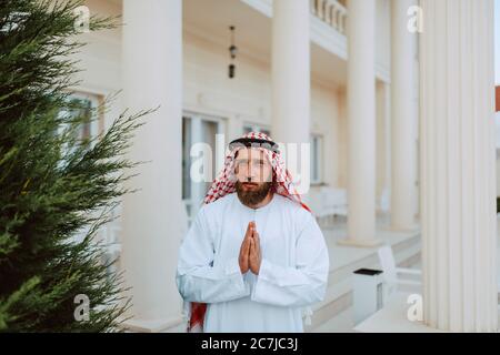 Bell'uomo musulmano arabo medio orientale in abiti tradizionali che si trova di fronte alla bella casa bianca moderna con palme ripiegate Foto Stock