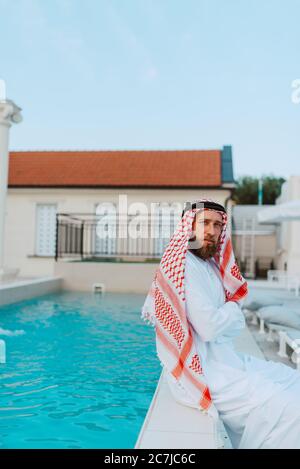 bell'uomo musulmano arabo medio-orientale in abiti tradizionali seduto sul bordo della piscina Foto Stock