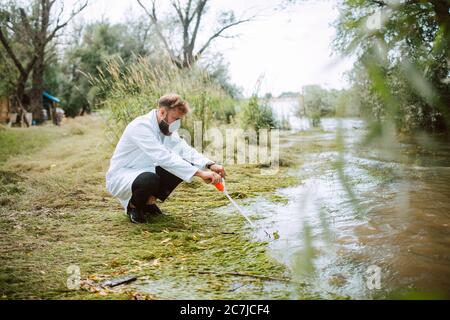 Biologo e ricercatore di scienziato caucasico maschile in tuta protettiva con maschera che preleva campioni d'acqua dal fiume inquinato. Foto Stock