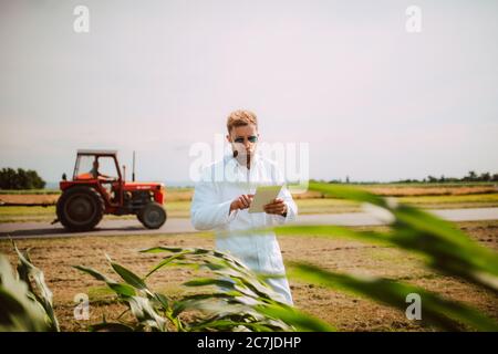 Agronomo tecnologo maschile con computer tablet nel campo del mais per il controllo della qualità e della crescita dei raccolti, mentre un trattore passa dietro di lui. Foto Stock