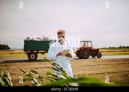 Agronomo tecnologo maschile con computer tablet nel campo del mais per il controllo della qualità e della crescita dei raccolti, mentre un trattore passa dietro di lui. Foto Stock